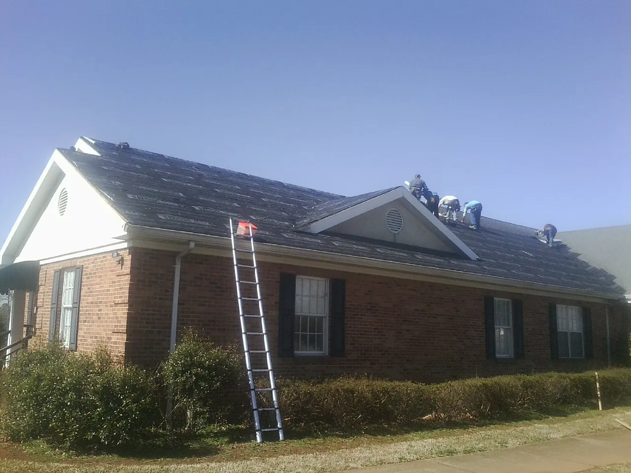 Crew tearing shingles off a brick ranch roof with a ladder set — Emergency Roof Repair work by our crew in Pink Hill