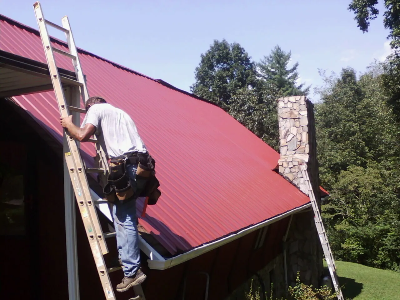 Roofer on a ladder beside a red metal roof and stone chimney — Soffit & Fascia Repair by Roofing Brothers in Pink Hill